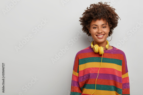 Energetic enthusiastic woman with curly crisp hair, listens music via headset, smiles broadly, being in high spirit, stands against white background with blank space, wears striped colorful jumper