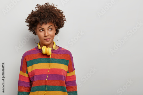 Photo of thoughtful curly haired woman looks aside with pensive expression, wears headphones and striped jumper, has plan in mind, poses over white background, blank space for your advertising content