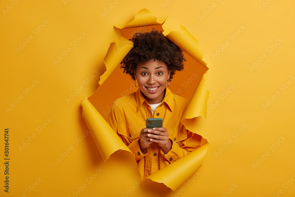 Happy curly haired student laughs at funny joke in social networks, smiles toothily, plays game online on modern cellphone, dressed in stylish outfit, stands in ripped hole background, yellow color - obrazy, fototapety, plakaty 