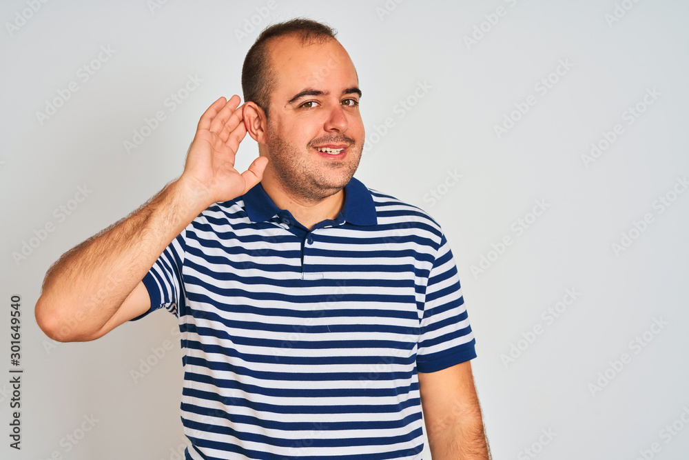 Young man wearing casual striped polo standing over isolated white background smiling with hand over ear listening an hearing to rumor or gossip. Deafness concept. Young man wearing casual striped polo standing over isolated white background smiling with hand over ear listening an hearing to rumor or gossip. Deafness concept.
