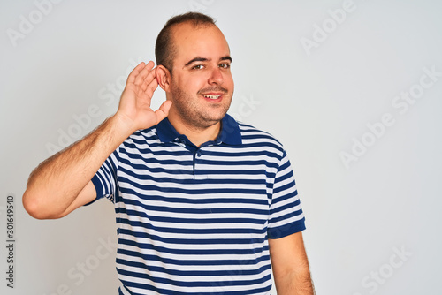 Young man wearing casual striped polo standing over isolated white background smiling with hand over ear listening an hearing to rumor or gossip. Deafness concept.