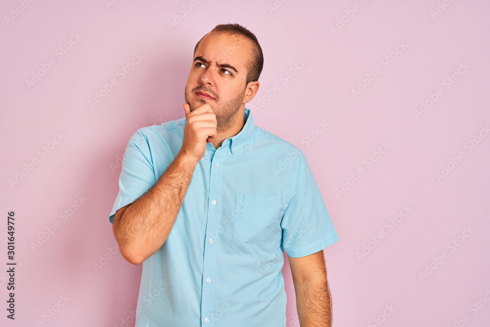 Young man wearing blue casual shirt standing over isolated pink background with hand on chin thinking about question, pensive expression. Smiling with thoughtful face. Doubt concept. Young man wearing blue casual shirt standing over isolated pink background with hand on chin thinking about question, pensive expression. Smiling with thoughtful face. Doubt concept.
