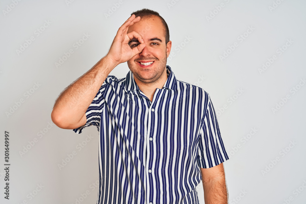 Young man wearing blue striped shirt standing over isolated white background doing ok gesture with hand smiling, eye looking through fingers with happy face. Young man wearing blue striped shirt standing over isolated white background doing ok gesture with hand smiling, eye looking through fingers with happy face.