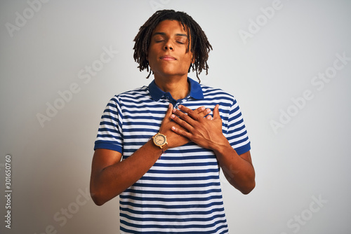 Afro man with dreadlocks wearing striped blue polo standing over isolated white background smiling with hands on chest with closed eyes and grateful gesture on face. Health concept.