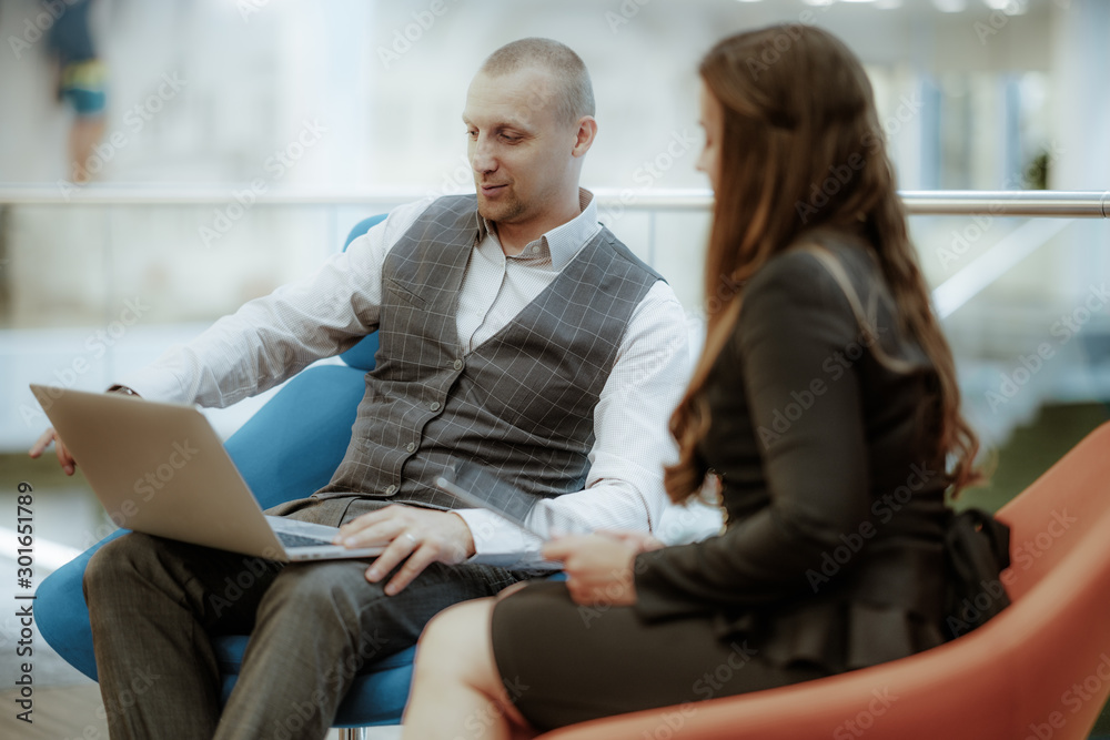 A handsome cheerful businessman is showing to his female colleague something on a screen of his laptop while both sitting on armchairs in an open space area of a contemporary office during a meeting