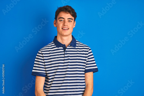 Teenager boy wearing casual t-shirt standing over blue isolated background looking away to side with smile on face, natural expression. Laughing confident.