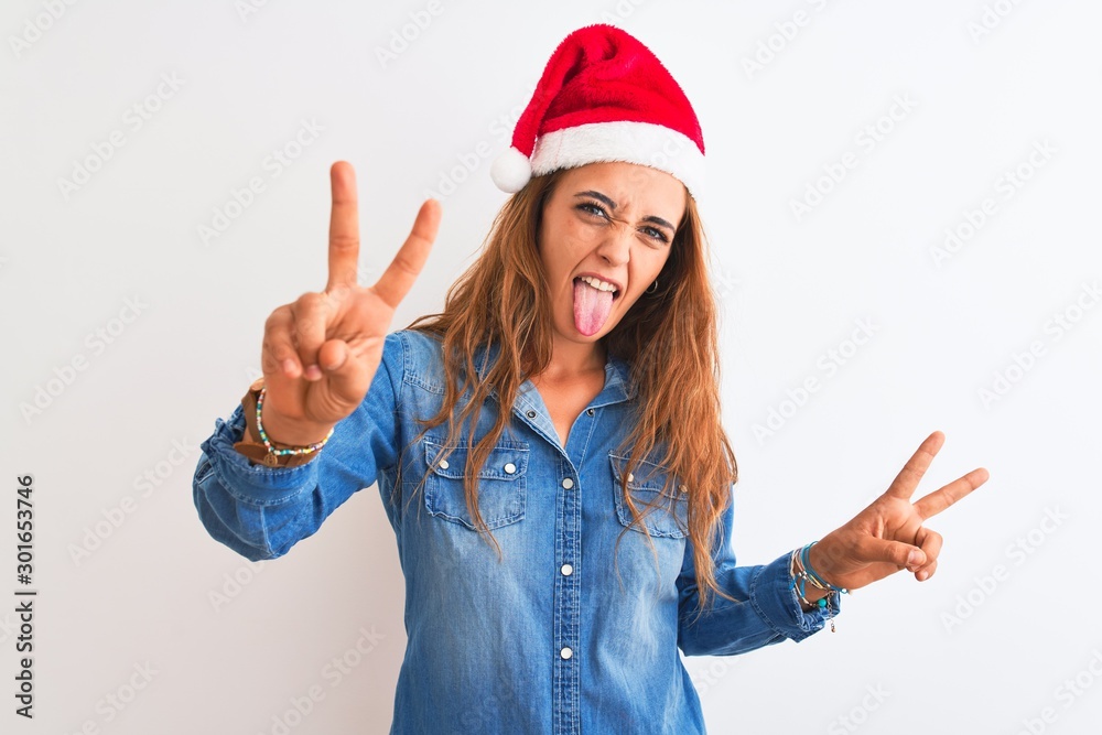 Young beautiful redhead woman wearing christmas hat over isolated background smiling with tongue out showing fingers of both hands doing victory sign. Number two. Young beautiful redhead woman wearing christmas hat over isolated background smiling with tongue out showing fingers of both hands doing victory sign. Number two.