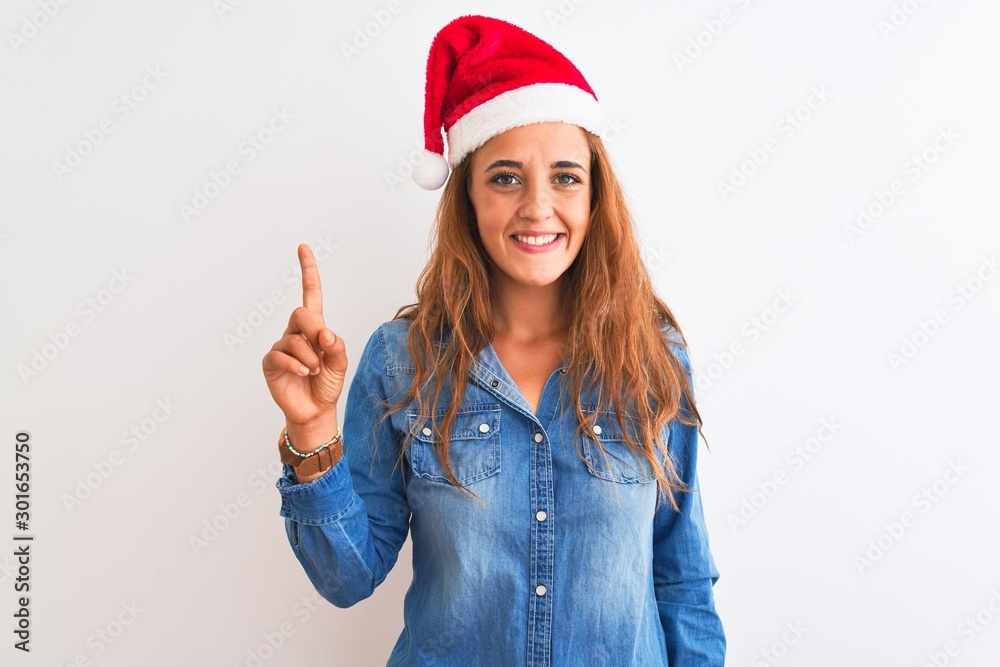 Young beautiful redhead woman wearing christmas hat over isolated background showing and pointing up with finger number one while smiling confident and happy. Young beautiful redhead woman wearing christmas hat over isolated background showing and pointing up with finger number one while smiling confident and happy.