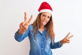 Young beautiful redhead woman wearing christmas hat over isolated background smiling with tongue out showing fingers of both hands doing victory sign. Number two.