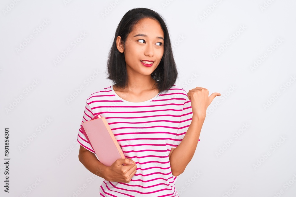 Young beautiful chinese woman holding books standing over isolated white background pointing and showing with thumb up to the side with happy face smiling Young beautiful chinese woman holding books standing over isolated white background pointing and showing with thumb up to the side with happy face smiling