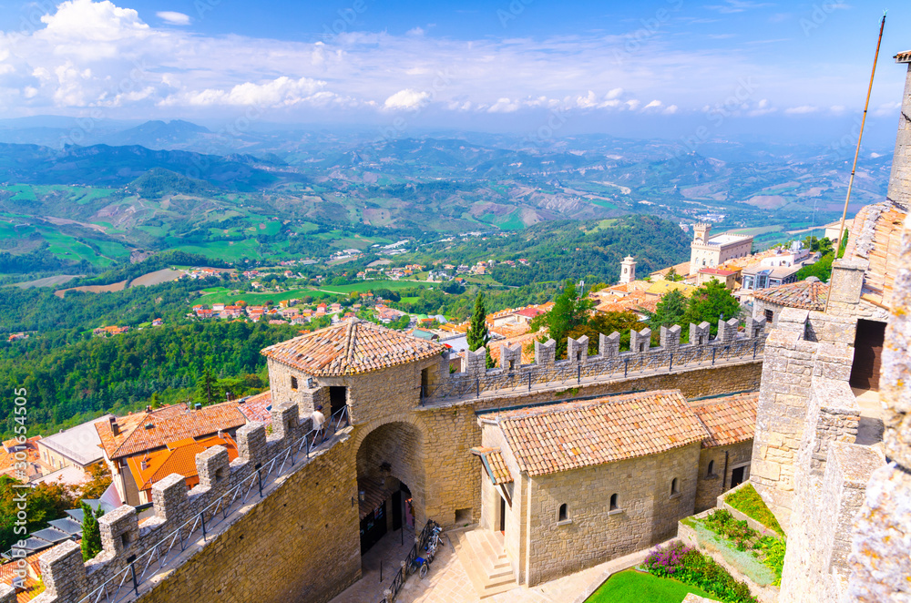 Aerial top panoramic view landscape with valley, green hills, fields, villages of Republic San Marino suburban district, blue sky white clouds background and stone fortress wall and tower with merlons