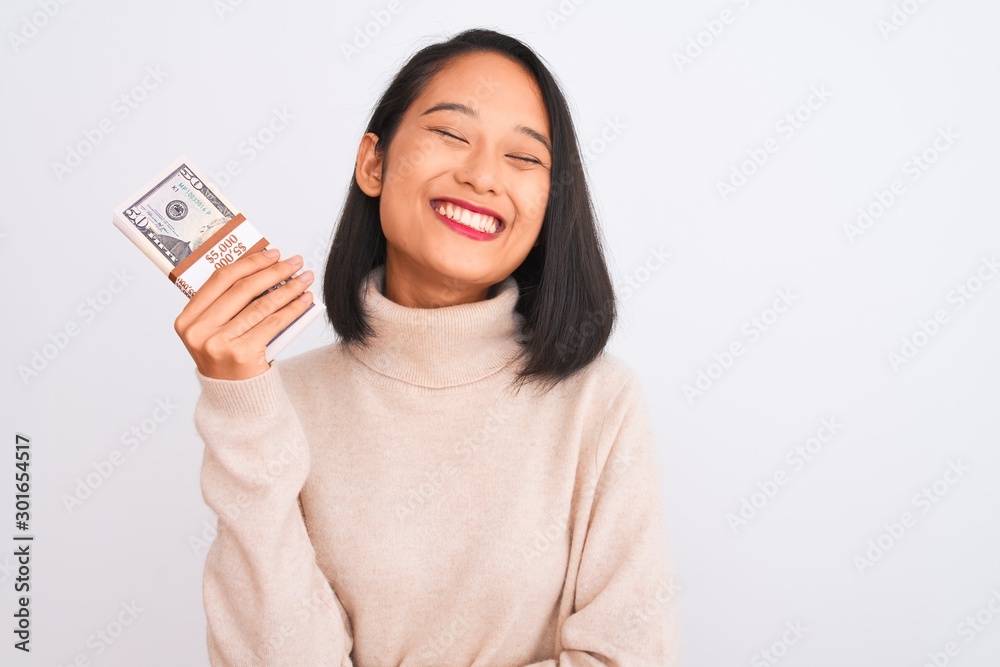 Young beautiful chinese woman holding dollars standing over isolated white background with a happy face standing and smiling with a confident smile showing teeth Young beautiful chinese woman holding dollars standing over isolated white background with a happy face standing and smiling with a confident smile showing teeth