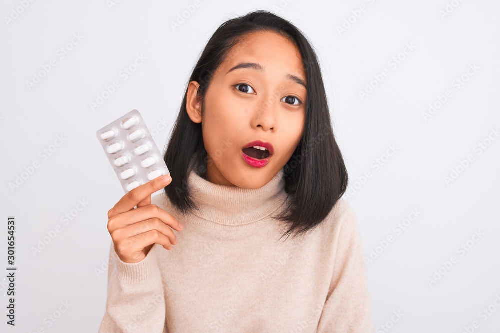 Young beautiful chinese woman holding pills standing over isolated white background scared in shock with a surprise face, afraid and excited with fear expression Young beautiful chinese woman holding pills standing over isolated white background scared in shock with a surprise face, afraid and excited with fear expression