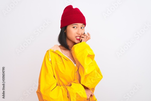 Young beautiful chinese woman wearing raincoat and wool cap over isolated white background looking stressed and nervous with hands on mouth biting nails. Anxiety problem. © Krakenimages.com