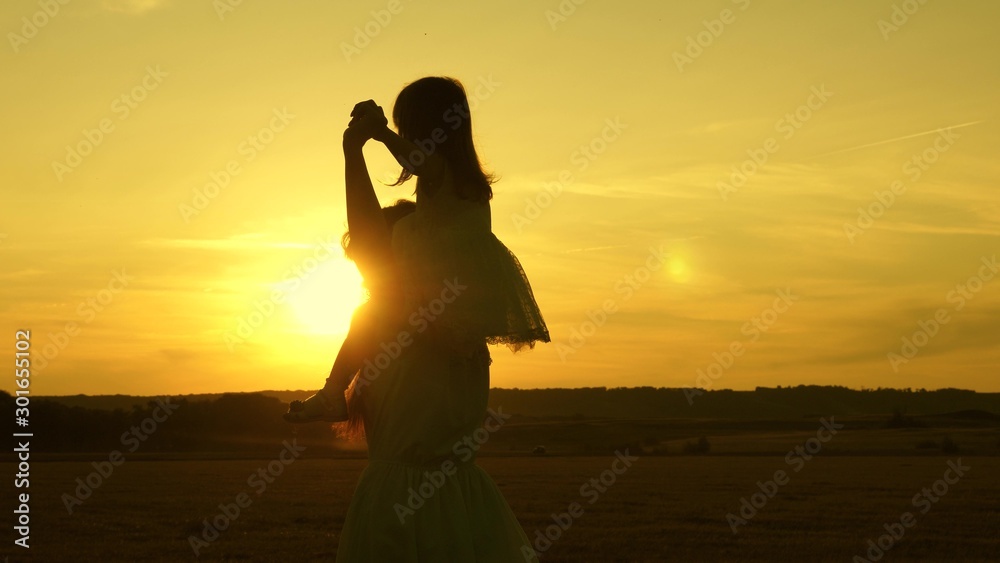 Happy mom and baby walk across the field at sunset and laugh. A little girl sits on her mother shoulders and walks together in the park in the sun and smiles. Family life concept. Slow motion.