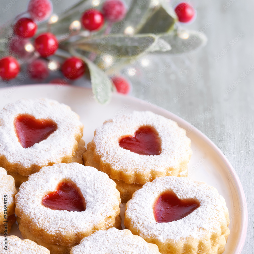 Close-up on traditional Christmas Linzer cookies, sandwich cookies  filled with strawberry jam on light table with Xmas decorations - festive lights and mistletoe with red berries.