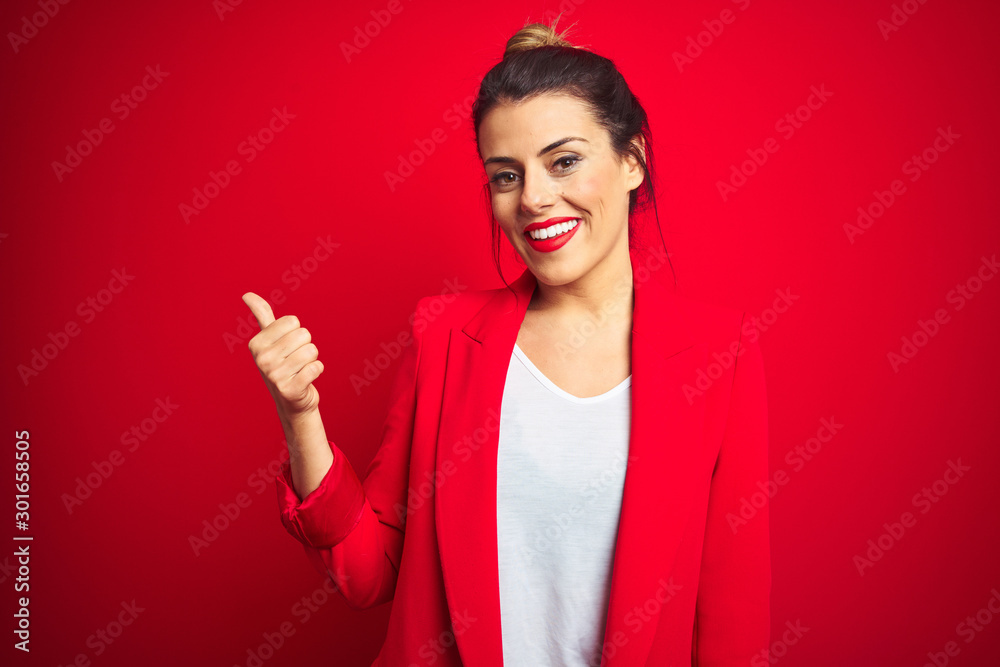 Young beautiful business woman standing over red isolated background smiling with happy face looking and pointing to the side with thumb up. Young beautiful business woman standing over red isolated background smiling with happy face looking and pointing to the side with thumb up.