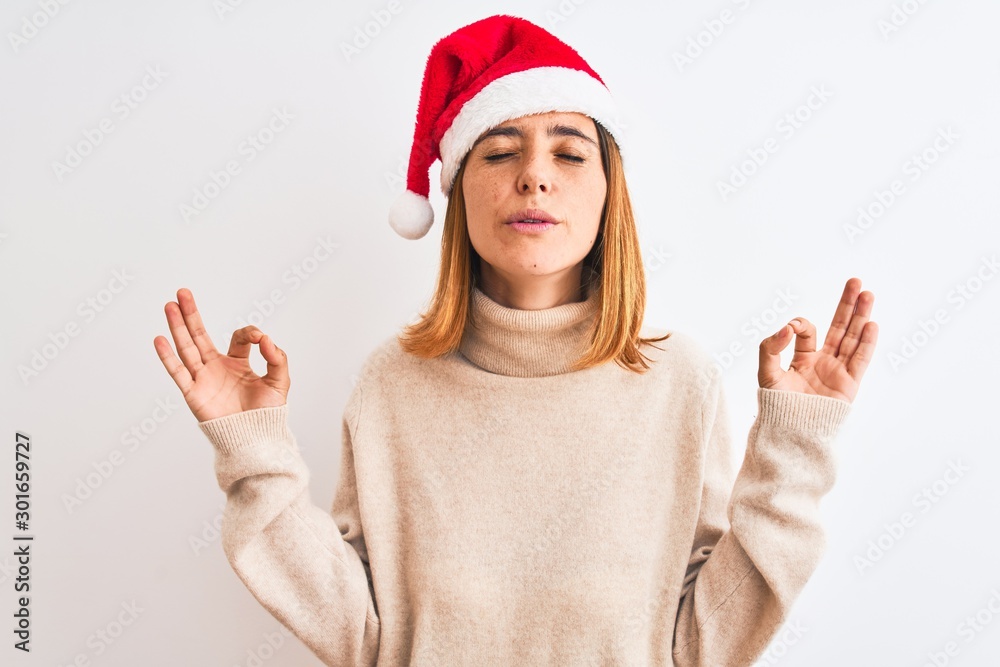 Beautiful redhead woman wearing christmas hat over isolated background relaxed and smiling with eyes closed doing meditation gesture with fingers. Yoga concept. Beautiful redhead woman wearing christmas hat over isolated background relaxed and smiling with eyes closed doing meditation gesture with fingers. Yoga concept.