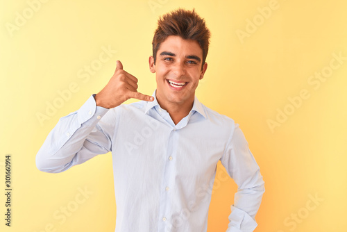 Young handsome businessman wearing elegant shirt over isolated yellow background smiling doing phone gesture with hand and fingers like talking on the telephone. Communicating concepts.