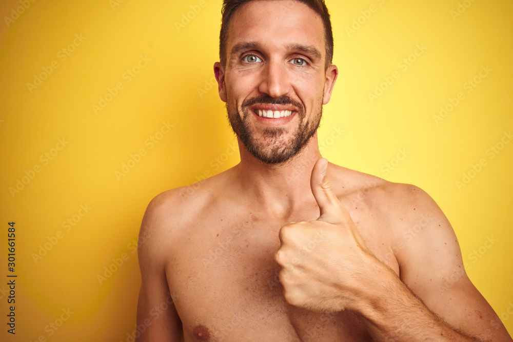 Fototapeta premium Sexy young shirtless man showing nude fitness chest over yellow isolated background doing happy thumbs up gesture with hand. Approving expression looking at the camera with showing success.