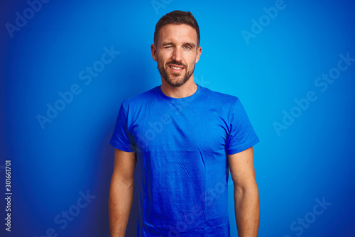 Young handsome man wearing casual t-shirt over blue isolated background winking looking at the camera with sexy expression, cheerful and happy face.
