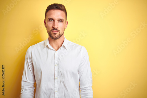 Young handsome man wearing elegant white shirt over yellow isolated background with serious expression on face. Simple and natural looking at the camera. © Krakenimages.com