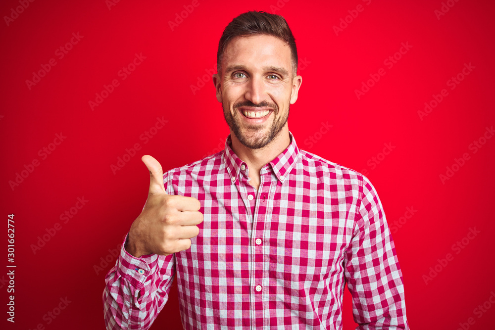 Young handsome man over red isolated background doing happy thumbs up gesture with hand. Approving expression looking at the camera showing success. Young handsome man over red isolated background doing happy thumbs up gesture with hand. Approving expression looking at the camera showing success.