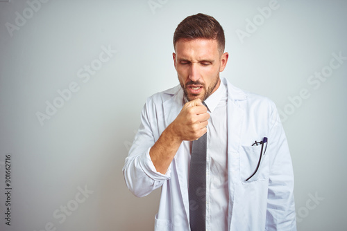 Young handsome doctor man wearing white profressional coat over isolated background feeling unwell and coughing as symptom for cold or bronchitis. Healthcare concept. photo