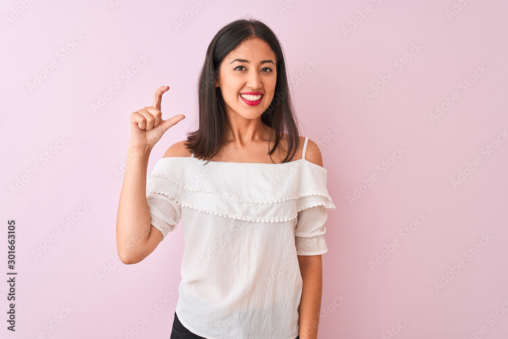 Beautiful chinese woman wearing white t-shirt standing over isolated pink background smiling and confident gesturing with hand doing small size sign with fingers looking and the camera Beautiful chinese woman wearing white t-shirt standing over isolated pink background smiling and confident gesturing with hand doing small size sign with fingers looking and the camera