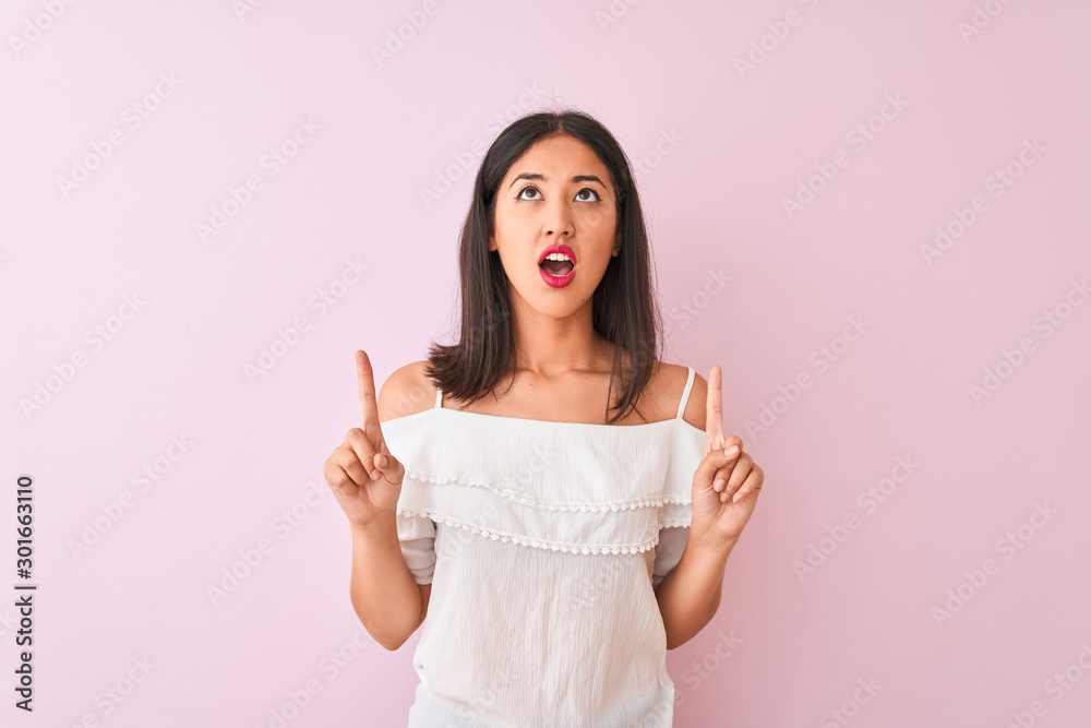 Beautiful chinese woman wearing white t-shirt standing over isolated pink background amazed and surprised looking up and pointing with fingers and raised arms. Beautiful chinese woman wearing white t-shirt standing over isolated pink background amazed and surprised looking up and pointing with fingers and raised arms.