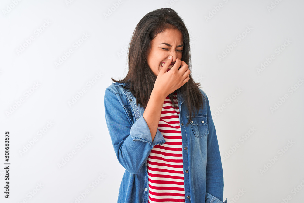 Young chinese woman wearing striped t-shirt and denim shirt over isolated white background smelling something stinky and disgusting, intolerable smell, holding breath with fingers on nose. Bad smell