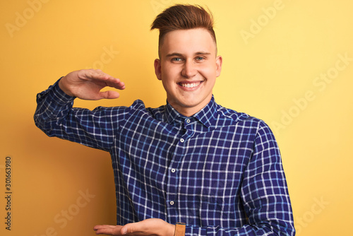 Young handsome man wearing casual shirt standing over isolated yellow background gesturing with hands showing big and large size sign, measure symbol. Smiling looking at the camera. Measuring concept.