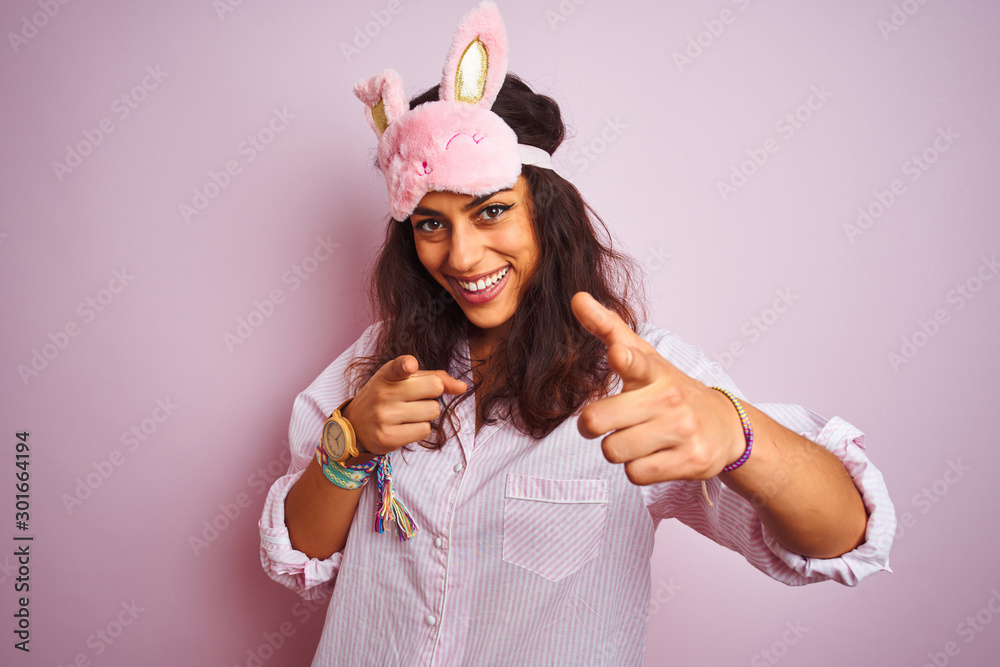 Young beautiful woman wearing pajama and sleep mask over isolated pink background pointing fingers to camera with happy and funny face. Good energy and vibes.