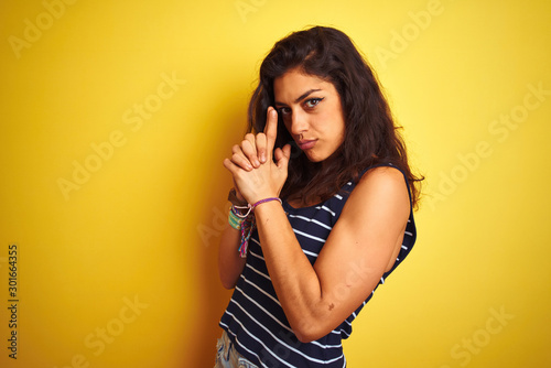 Young beautiful woman wearing striped t-shirt standing over isolated yellow background Holding symbolic gun with hand gesture, playing killing shooting weapons, angry face