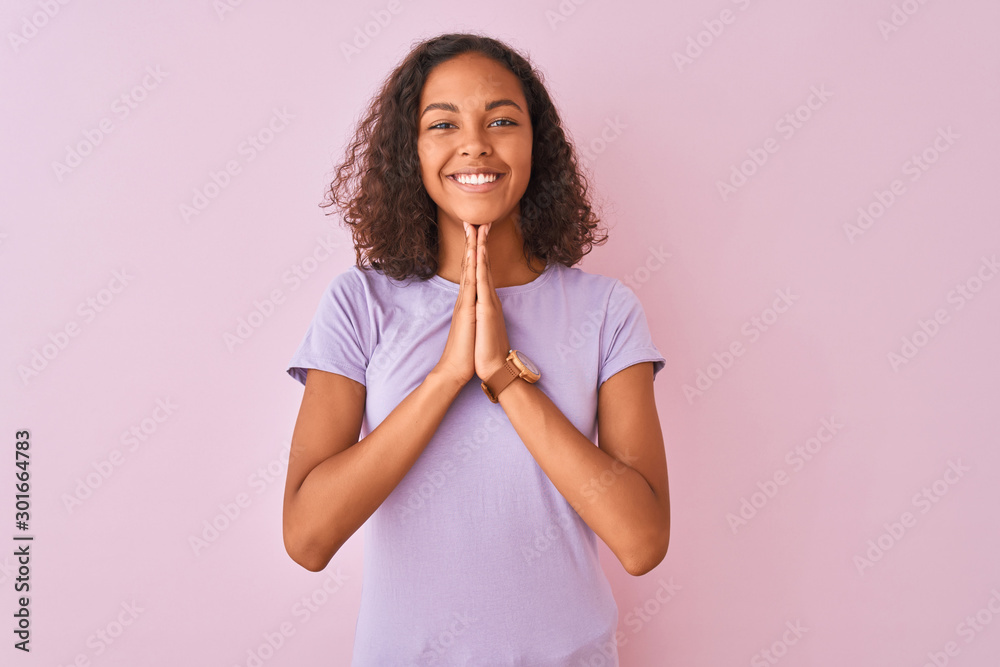 Young brazilian woman wearing t-shirt standing over isolated pink background praying with hands together asking for forgiveness smiling confident. - obrazy, fototapety, plakaty 
