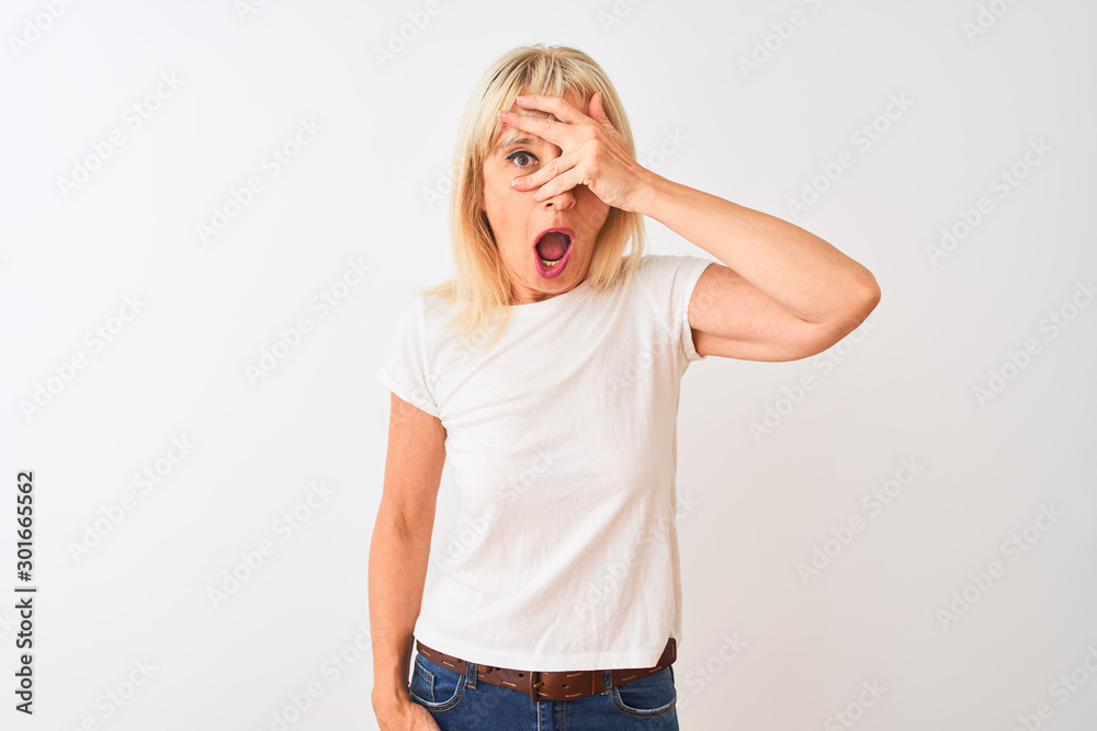 Middle age woman wearing casual t-shirt standing over isolated white background peeking in shock covering face and eyes with hand, looking through fingers with embarrassed expression. Middle age woman wearing casual t-shirt standing over isolated white background peeking in shock covering face and eyes with hand, looking through fingers with embarrassed expression.