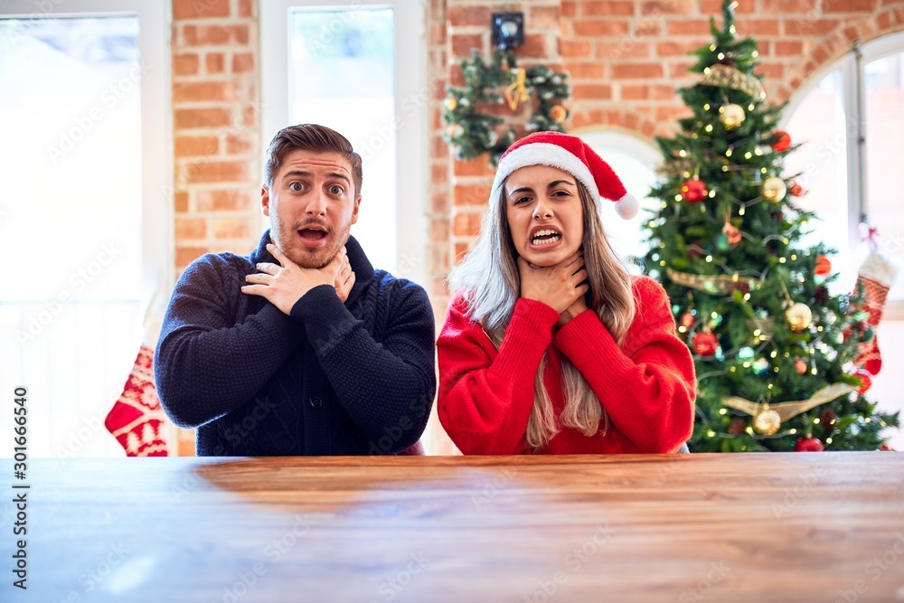 Young couple wearing santa claus hat sitting on chair and table around christmas tree at home shouting and suffocate because painful strangle. Health problem. Asphyxiate and suicide concept. Young couple wearing santa claus hat sitting on chair and table around christmas tree at home shouting and suffocate because painful strangle. Health problem. Asphyxiate and suicide concept.