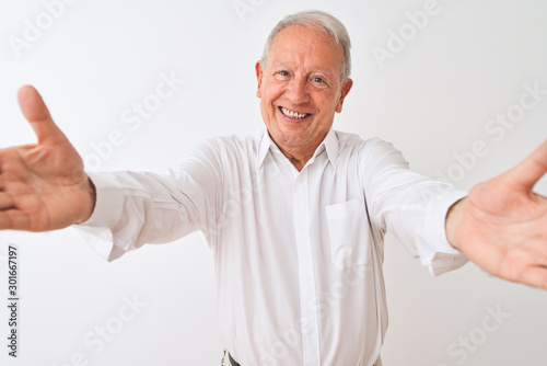 Senior grey-haired man wearing elegant shirt standing over isolated white background looking at the camera smiling with open arms for hug. Cheerful expression embracing happiness.