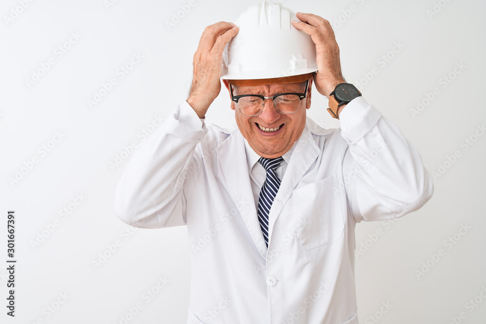 Senior grey-haired engineer man wearing coat and helmet over isolated white background suffering from headache desperate and stressed because pain and migraine. Hands on head. Senior grey-haired engineer man wearing coat and helmet over isolated white background suffering from headache desperate and stressed because pain and migraine. Hands on head.
