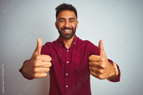 Young indian man wearing red elegant shirt standing over isolated grey background success sign doing positive gesture with hand, thumbs up smiling and happy. Cheerful expression and winner gesture. photo