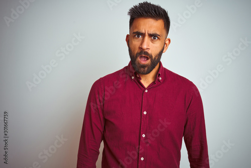 Young indian man wearing red elegant shirt standing over isolated grey background In shock face, looking skeptical and sarcastic, surprised with open mouth