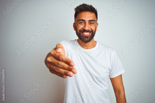 Young indian man wearing t-shirt standing over isolated white background smiling friendly offering handshake as greeting and welcoming. Successful business.