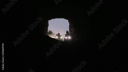 Row of several extreme quadrocycles switched on headlights and with men wearing helmets are actively riding and moving around inside of huge empty dark cave. photo