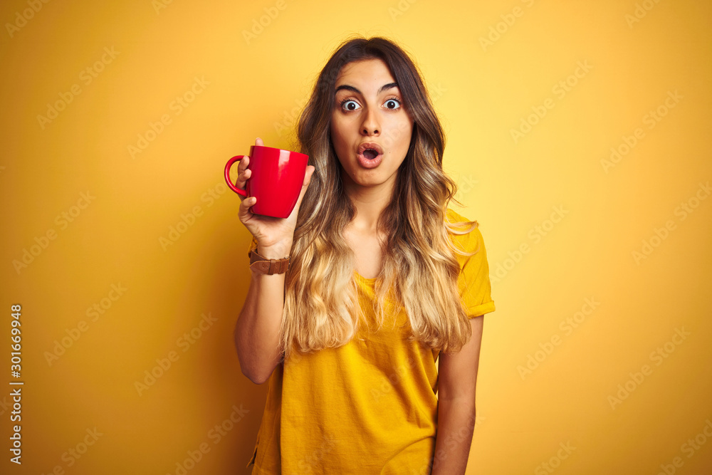 Young beautiful woman holding  red cup of coffee over yellow isolated background scared in shock with a surprise face, afraid and excited with fear expression