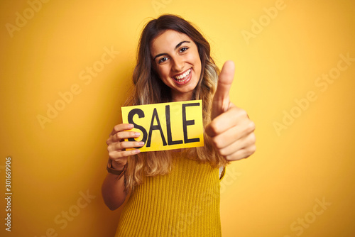 Young beautiful woman holding sale poster over yellow isolated background happy with big smile doing ok sign, thumb up with fingers, excellent sign