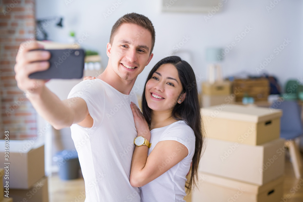 Young beautiful couple standing using smartphone to take a selfie at new home around cardboard boxes Young beautiful couple standing using smartphone to take a selfie at new home around cardboard boxes