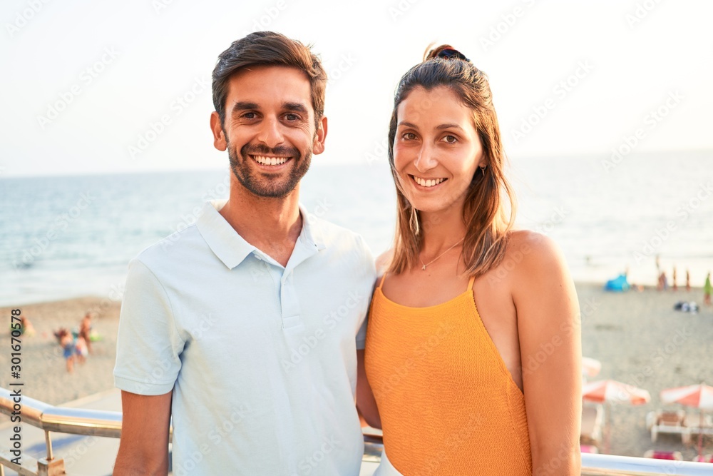 Young beautiful couple on vacation smiling happy and confident. Standing with smile on face hugging at the beach Young beautiful couple on vacation smiling happy and confident. Standing with smile on face hugging at the beach