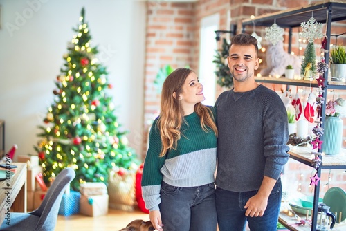 Young beautiful couple smiling happy and confident. Standing and hugging around christmas tree at home