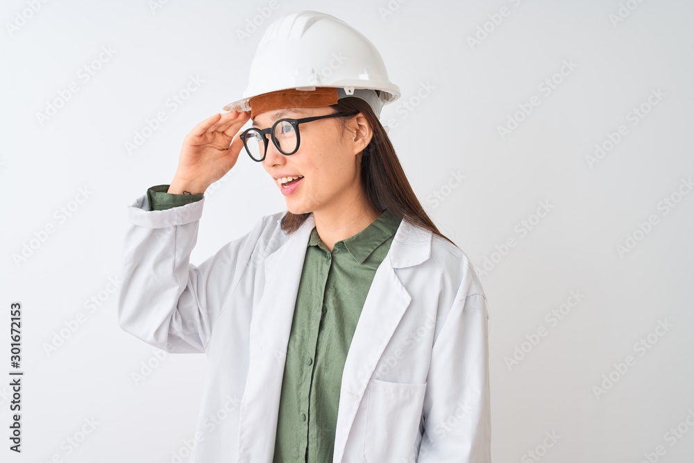 Young chinese engineer woman wearing coat helmet glasses over isolated white background very happy and smiling looking far away with hand over head. Searching concept. Young chinese engineer woman wearing coat helmet glasses over isolated white background very happy and smiling looking far away with hand over head. Searching concept.