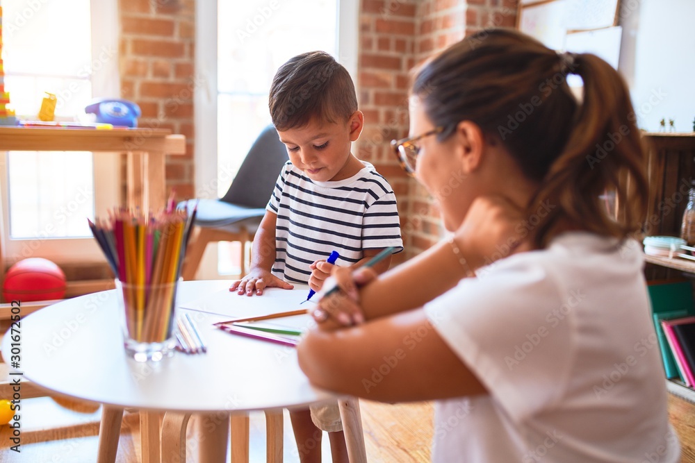 Beautiful teacher and toddler boy drawing draw using colored pencils at kindergarten Beautiful teacher and toddler boy drawing draw using colored pencils at kindergarten